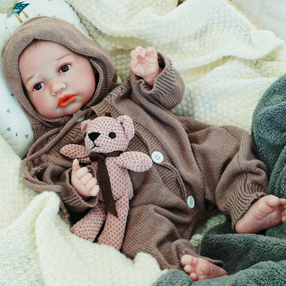 The reborn baby doll with an attentive gaze, dressed in a brown knit outfit, clutching a soft teddy bear, resting on a soft blanket.