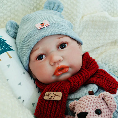 The reborn baby doll gazing upward, dressed warmly in a gray outfit with a red scarf and hat, holding a teddy bear.