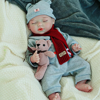 A reborn baby doll sitting up, wearing a gray outfit with a red scarf and hat, holding a pink teddy bear.