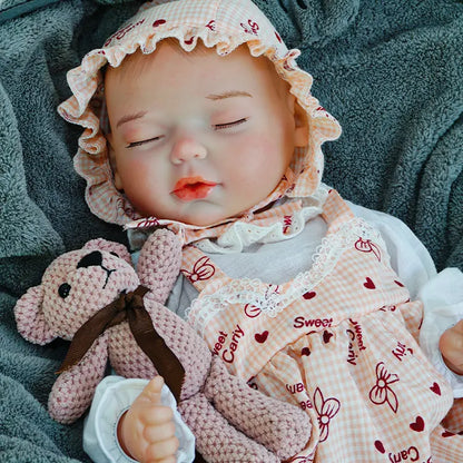 A reborn baby doll resting on a dark blanket, wearing a bonnet, holding a pink teddy bear.