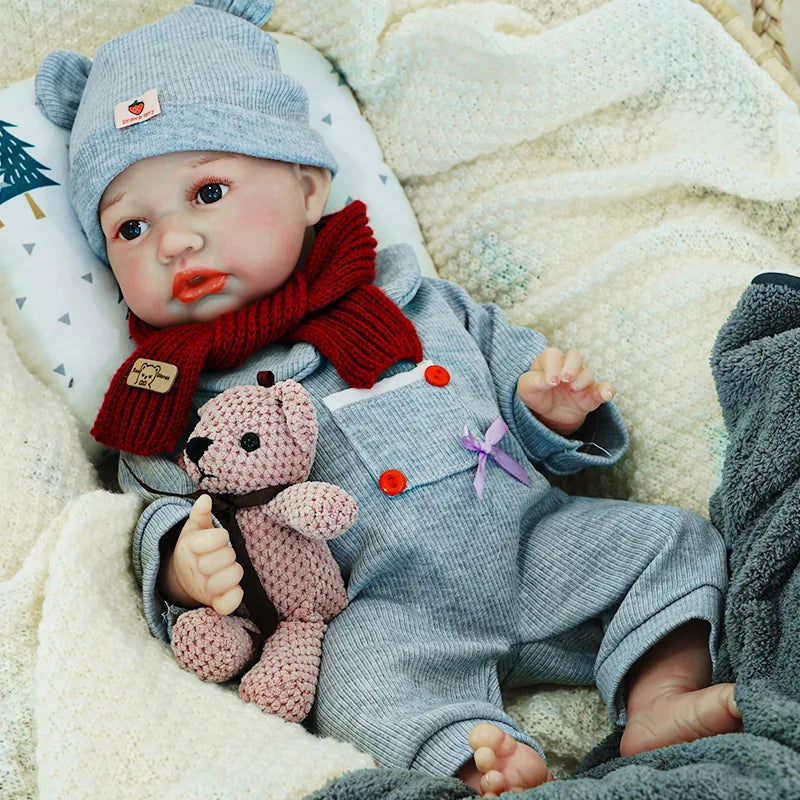 The reborn baby doll resting comfortably, dressed in a gray onesie with a red scarf, cuddling its teddy bear.