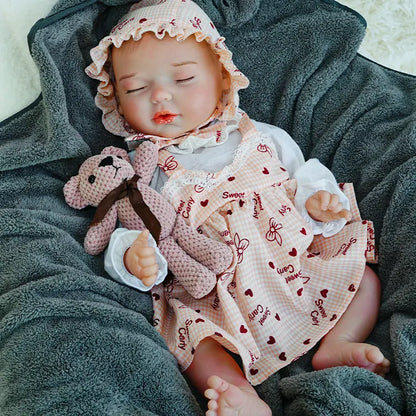 A reborn baby doll lying on a dark blanket, eyes closed, holding a pink teddy bear.