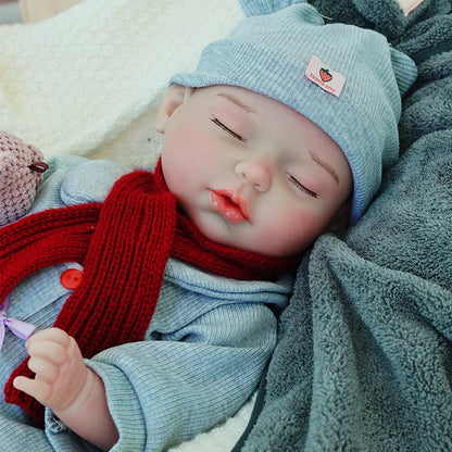 A reborn baby doll peacefully sleeping, dressed in a gray outfit with a red scarf, resting on a light blanket, holding a teddy bear.