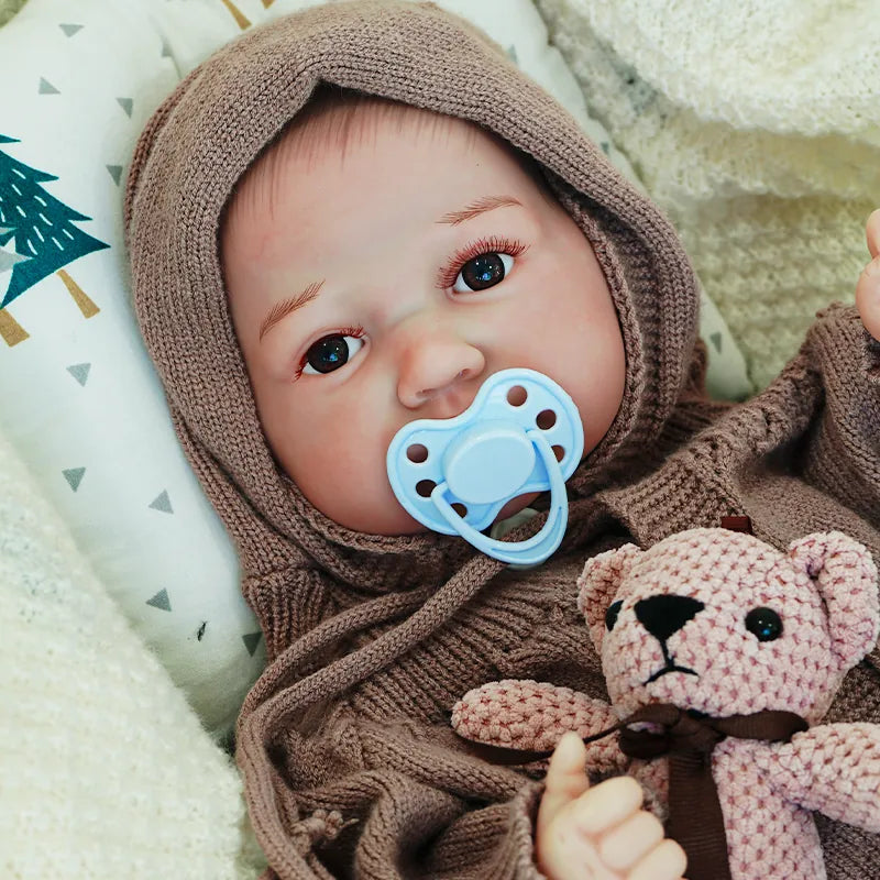 The reborn baby doll with a pacifier in its mouth, dressed in a brown knit onesie, holding a knitted teddy bear, looking peacefully at ease.