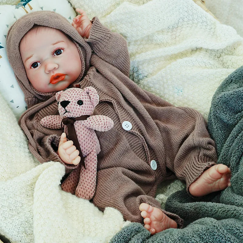 The reborn baby doll resting, dressed in a brown hooded onesie, holding a knitted teddy bear, with a lifelike and calm expression.