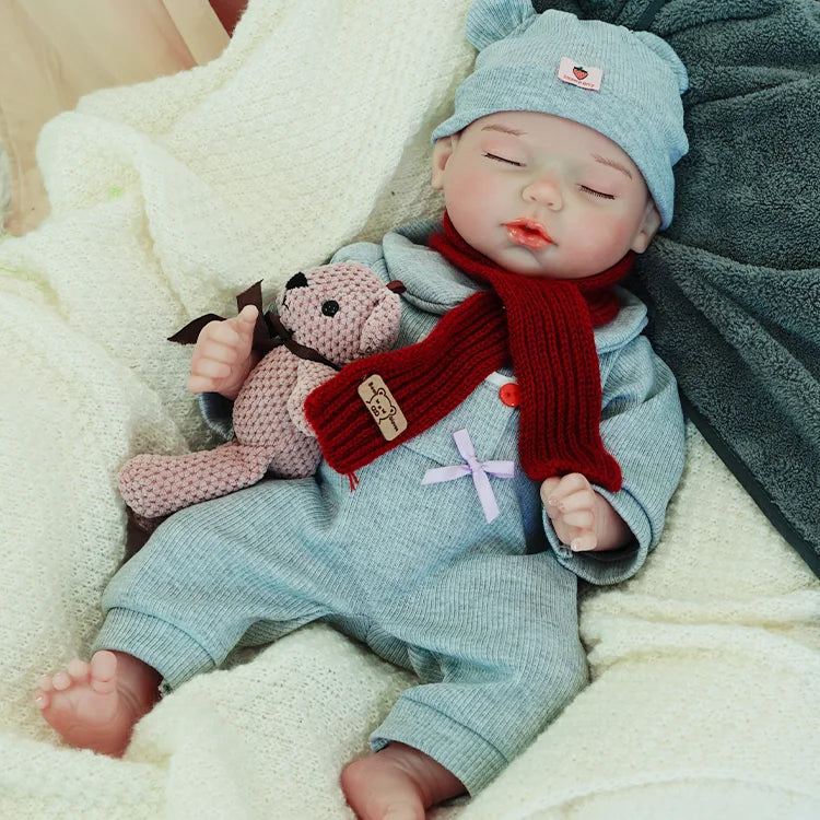 A reborn baby doll dressed in a gray outfit and hat, lying on a light blanket with a teddy bear, eyes closed.