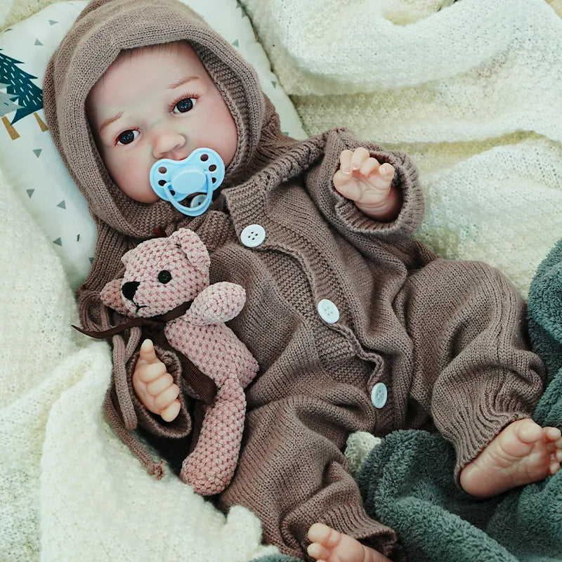 The reborn baby doll sitting, with a pacifier, dressed in a brown knit outfit, holding a soft teddy bear, offering a realistic and comforting presence.