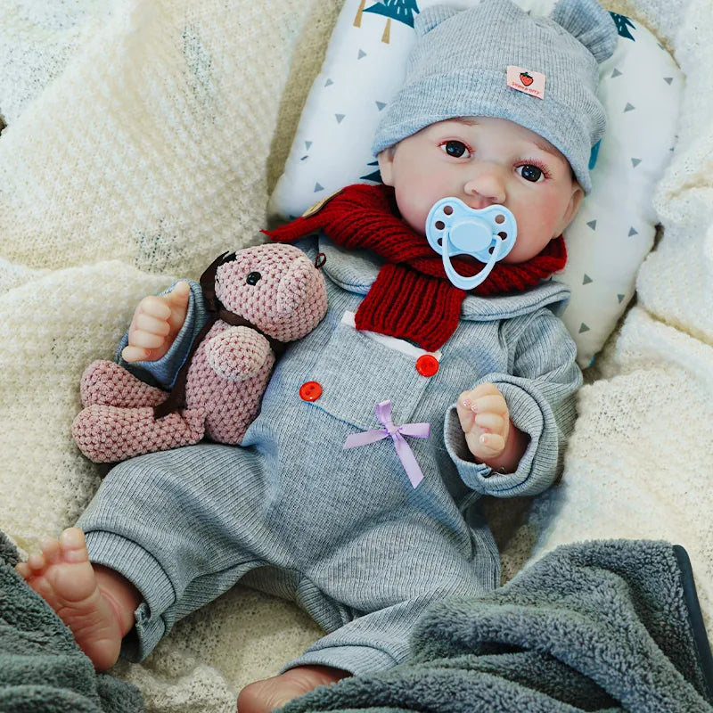 The reborn baby doll with a pacifier, dressed in a warm gray and red outfit, lying on a soft blanket with a teddy bear.