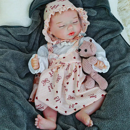 A reborn baby doll sitting up, holding a pink teddy bear, on a dark blanket.