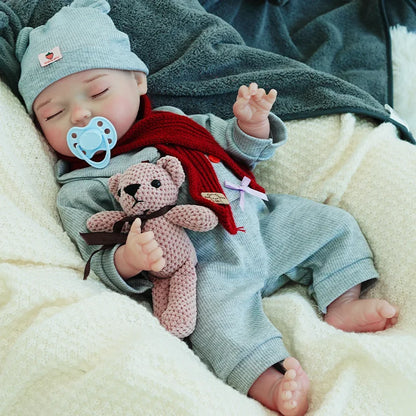 A reborn baby doll with a pacifier, dressed in a gray outfit and hat, lying on a light blanket, holding a teddy bear, arms raised.