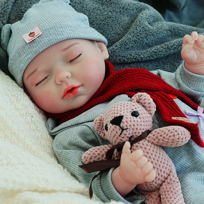 A reborn baby doll sleeping peacefully, dressed in a gray outfit with a red scarf, cuddling a teddy bear on a light blanket.