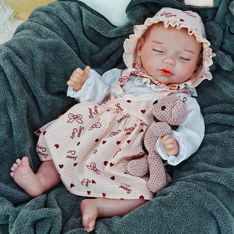 A reborn baby doll sitting up, holding a pink teddy bear, with a dark blanket