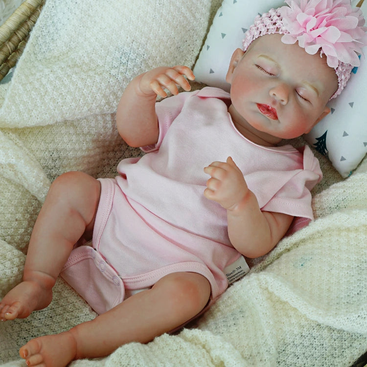 Reborn baby doll lying with eyes closed in a pink onesie.