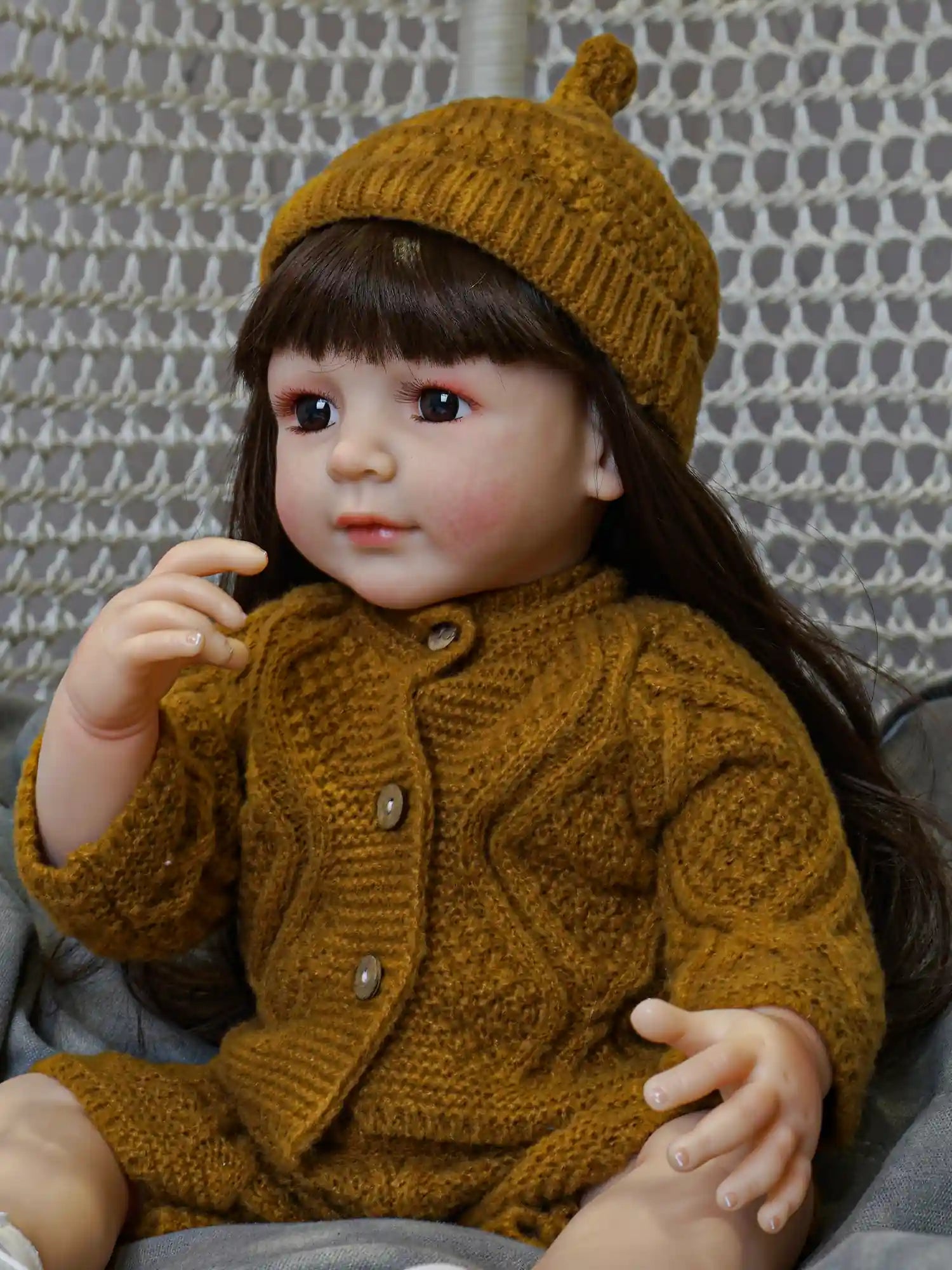 Collectible doll with a serene expression, featuring long brown hair and wearing seasonal mustard knitwear, positioned cozily with white shoes on a gray cushion.