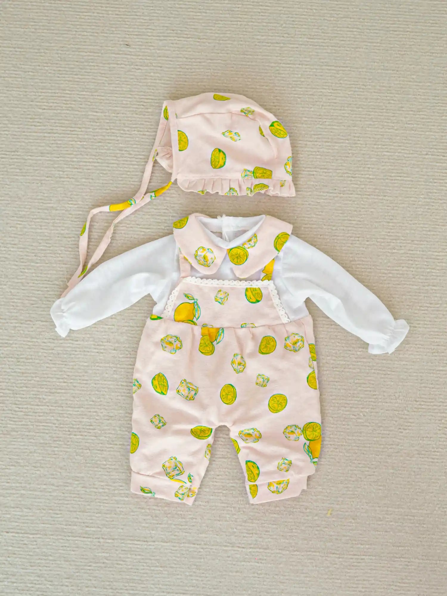 A reborn doll outfit displayed on a neutral background, featuring a pink strap overalls with a lemon print, accompanied by a matching bonnet and a white long-sleeved undershirt.