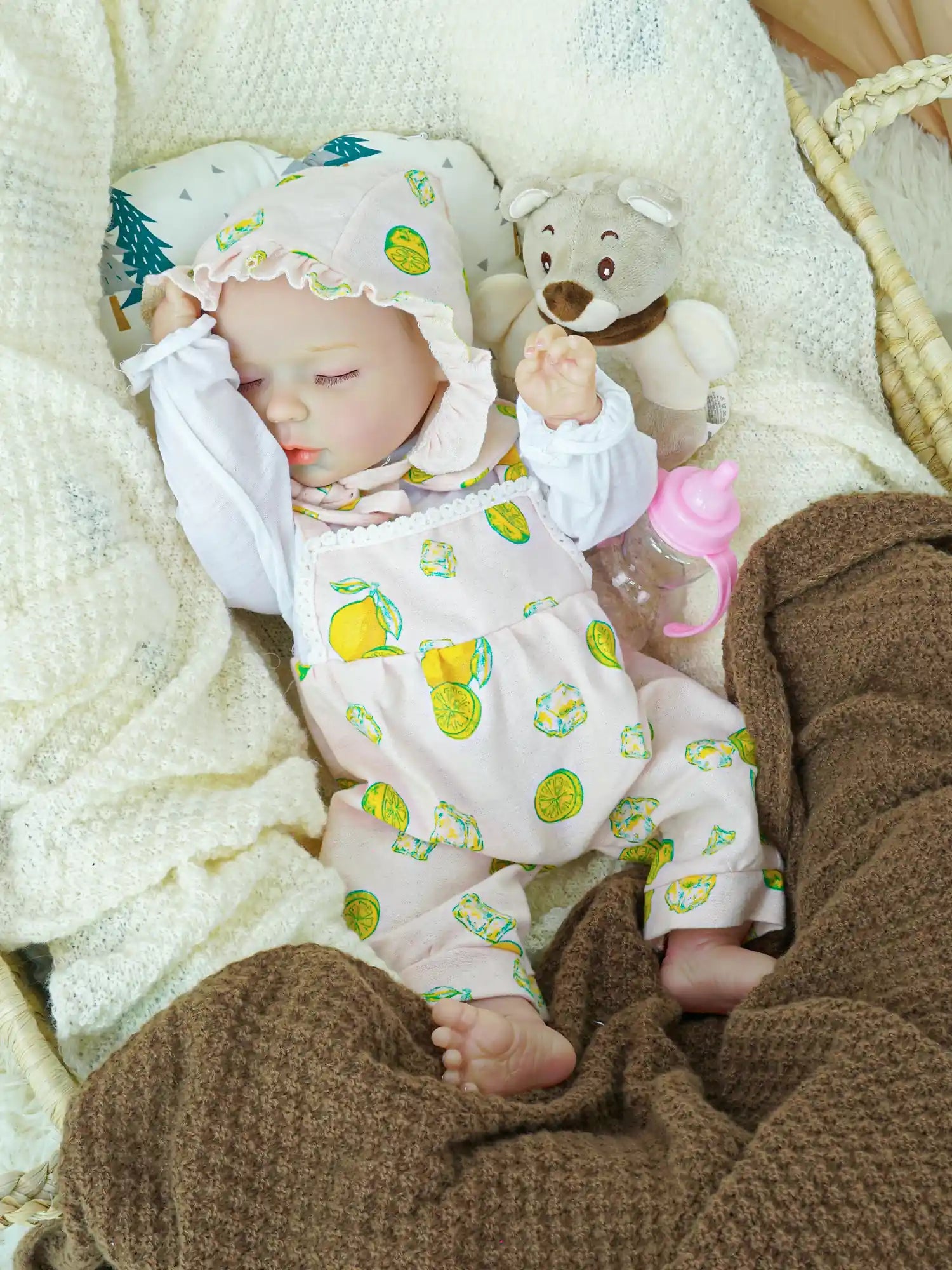 Close-up of a reborn baby doll in a pink lemon-print bonnet and outfit, resting in a cozy basket. The doll is surrounded by soft white and brown blankets, accompanied by a plush teddy bear and a baby bottle.