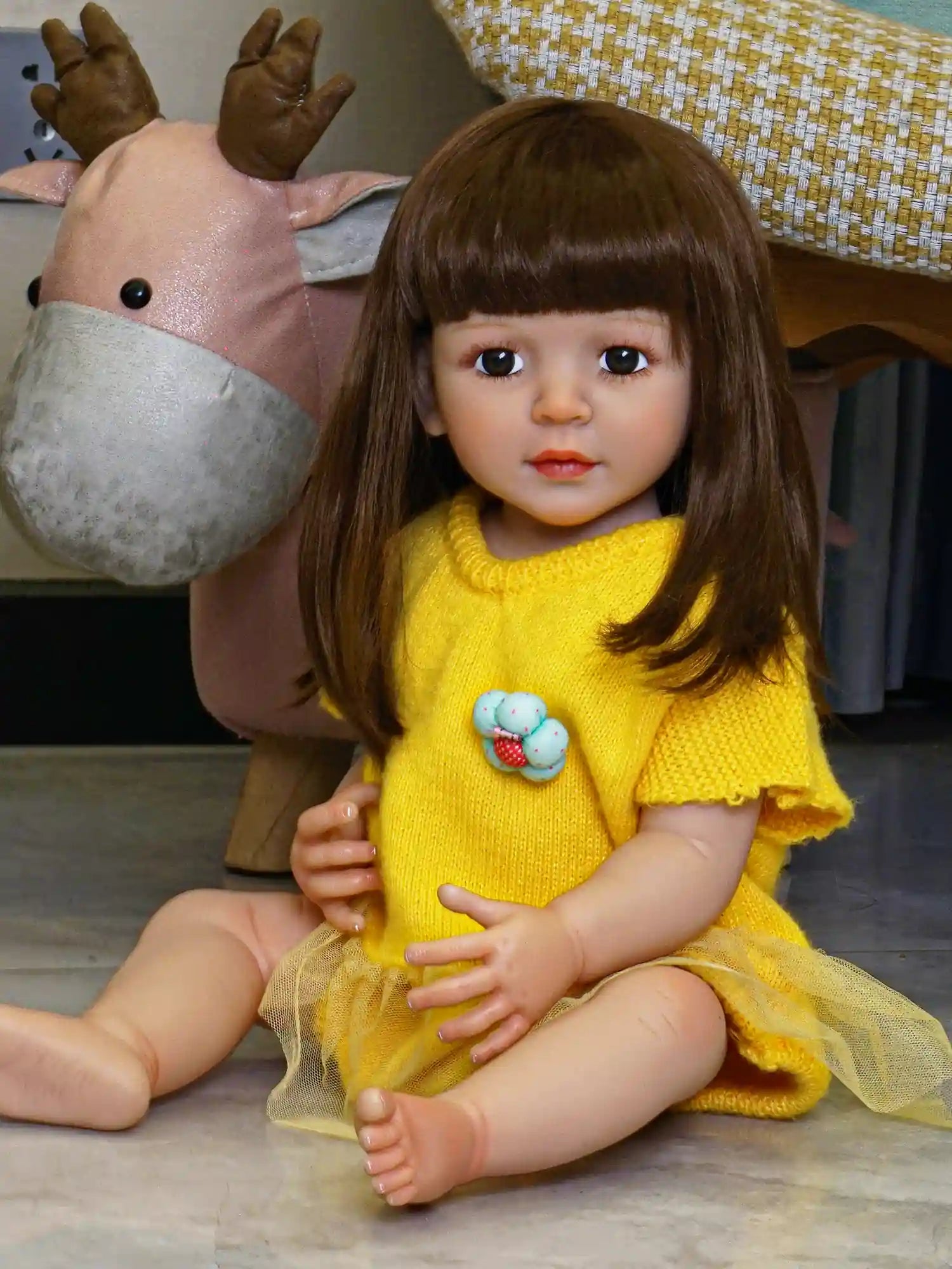 Artisanal doll with a serene countenance and soft brown hair, attired in a knit yellow garment with a flouncy skirt, in a cozy indoor setting with plush toys and decorative pillows.