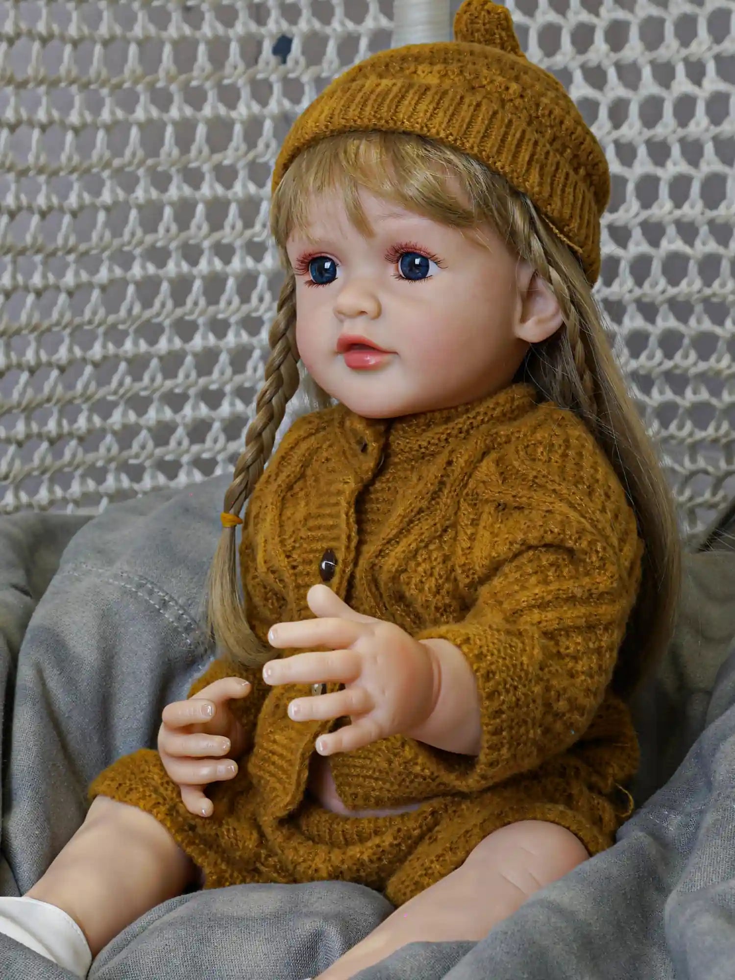A doll capturing the innocence of a toddler, with braided blonde hair and blue eyes, cozy in a textured mustard knitwear set against a soft grey surface.