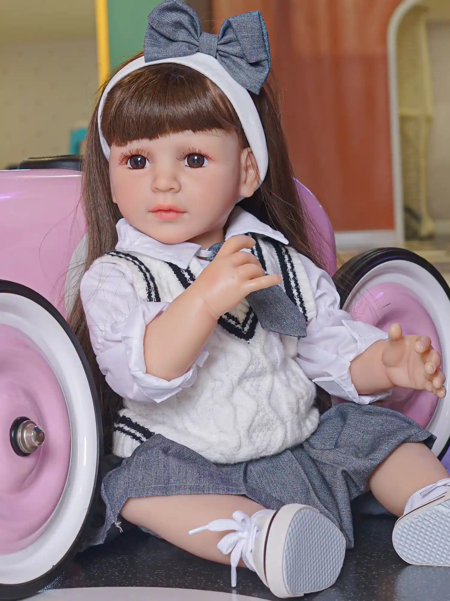 Realistic toddler doll in a seated position, with a dark hair bow and a traditional-looking school uniform, set against the backdrop of a children's play area.
