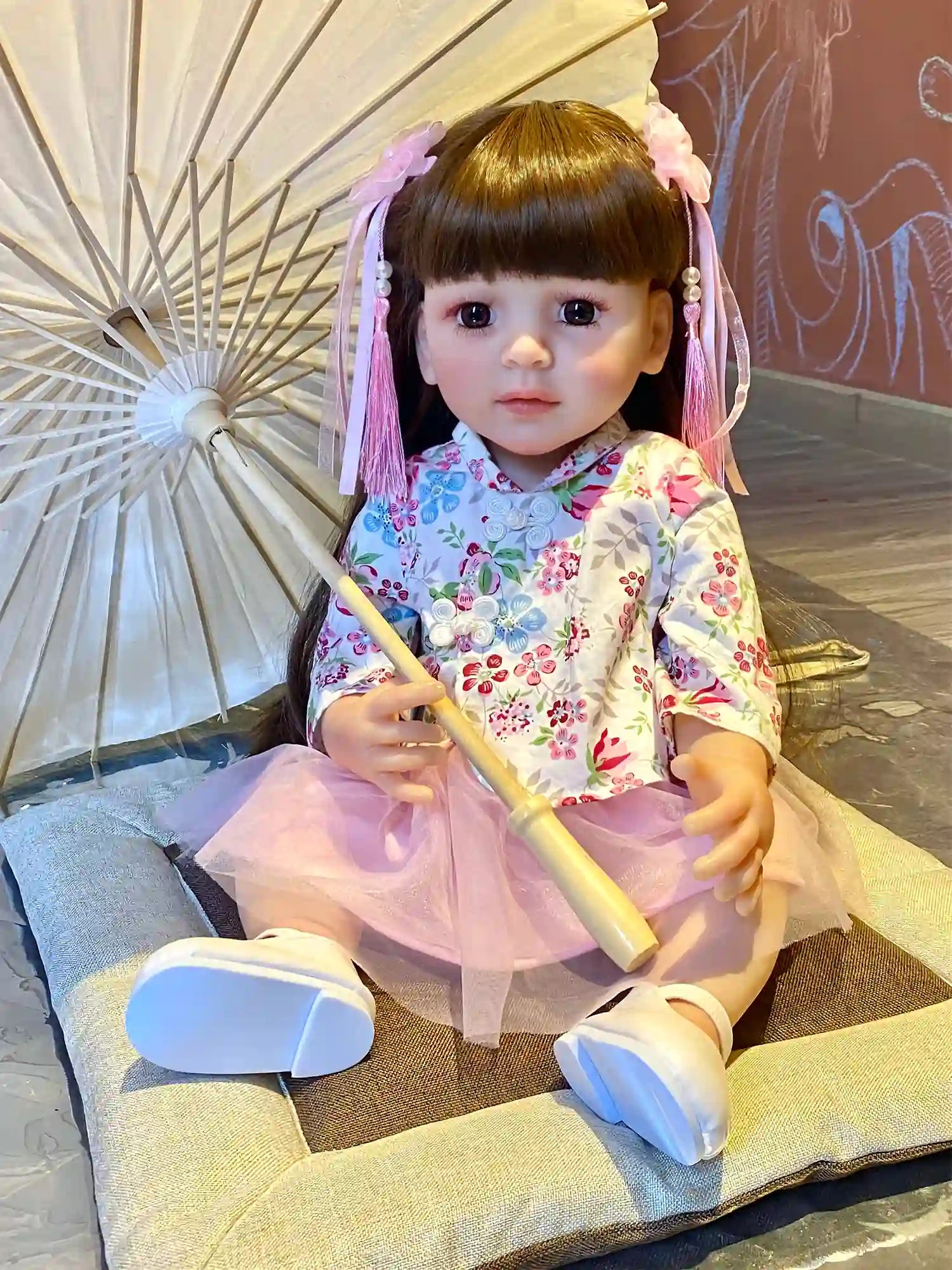 Toddler doll in a seated position, wearing a pink and white floral kimono dress and pink tulle skirt, holding a pink ribbon, with a playful expression.