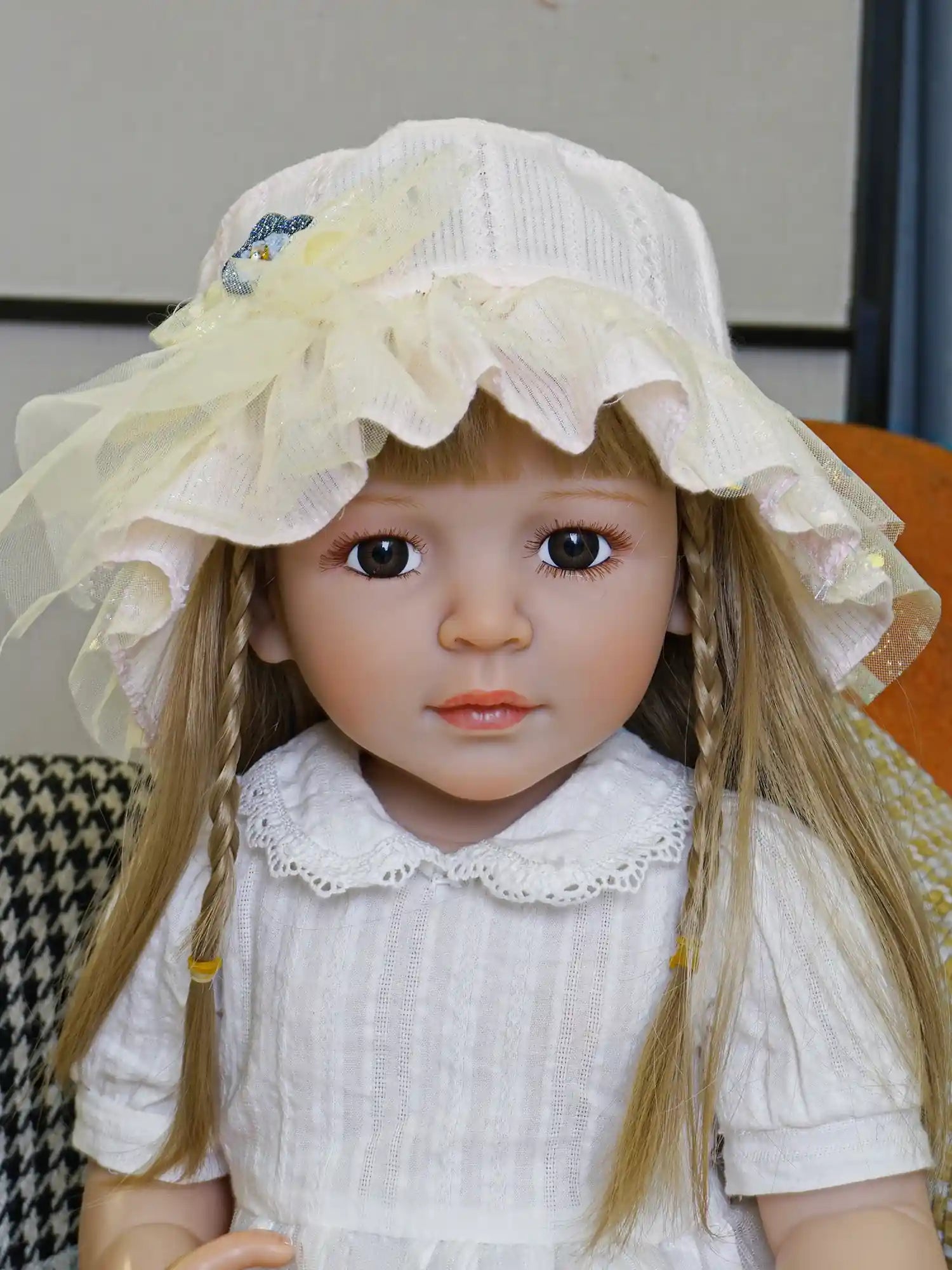 Realistic doll featuring braids and a yellow sunhat, posed on a chair with vibrant geometric cushions in the background.