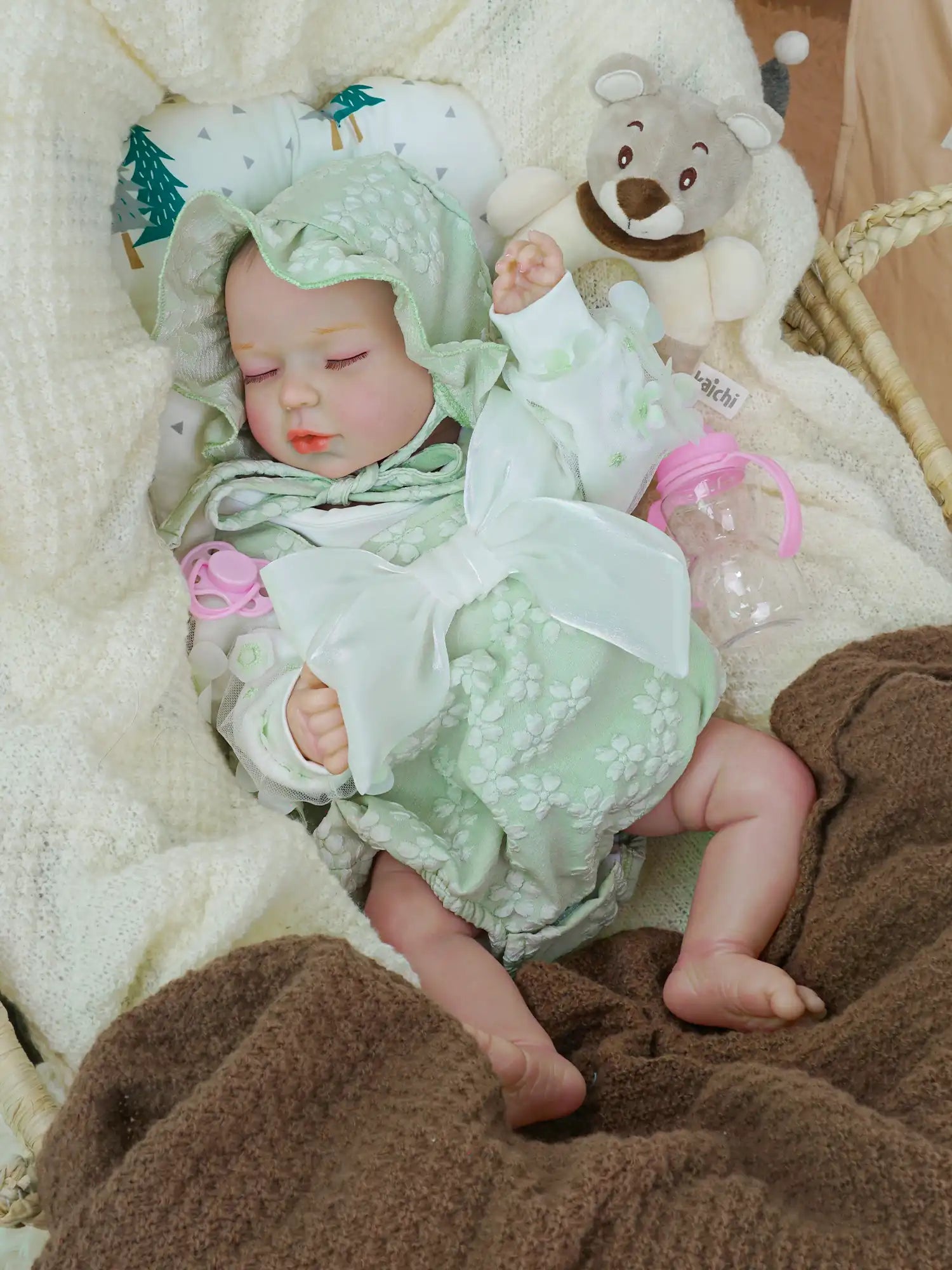 A serene reborn baby doll wrapped in a pale green floral romper and matching hood, peacefully sleeping in a wicker basket, nestled in a white knitted blanket, with a plush teddy bear and a pink bottle nearby.