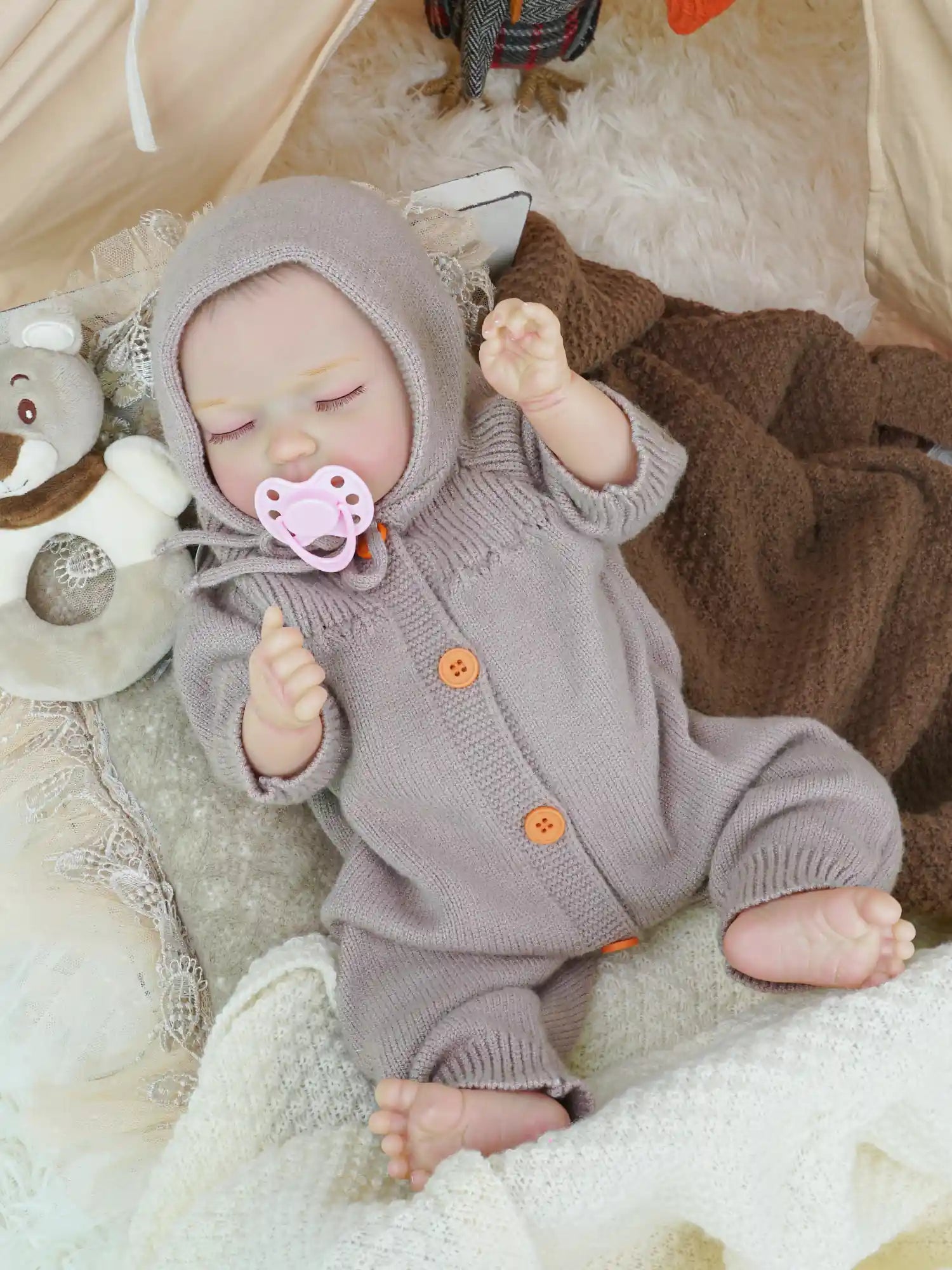 A reborn doll toy dressed in a lilac knitted outfit with a matching hood, laying on a soft white blanket, surrounded by plush toys and a tent background.