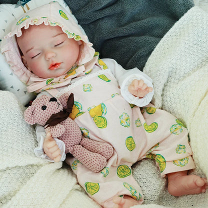 Close-up of a reborn baby doll’s face, dressed in a light green outfit, with a knitted bear beside it.