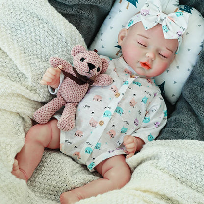 A sleeping reborn baby doll wearing a light onesie with blue patterns and a large bow headband, holding a knitted teddy bear.