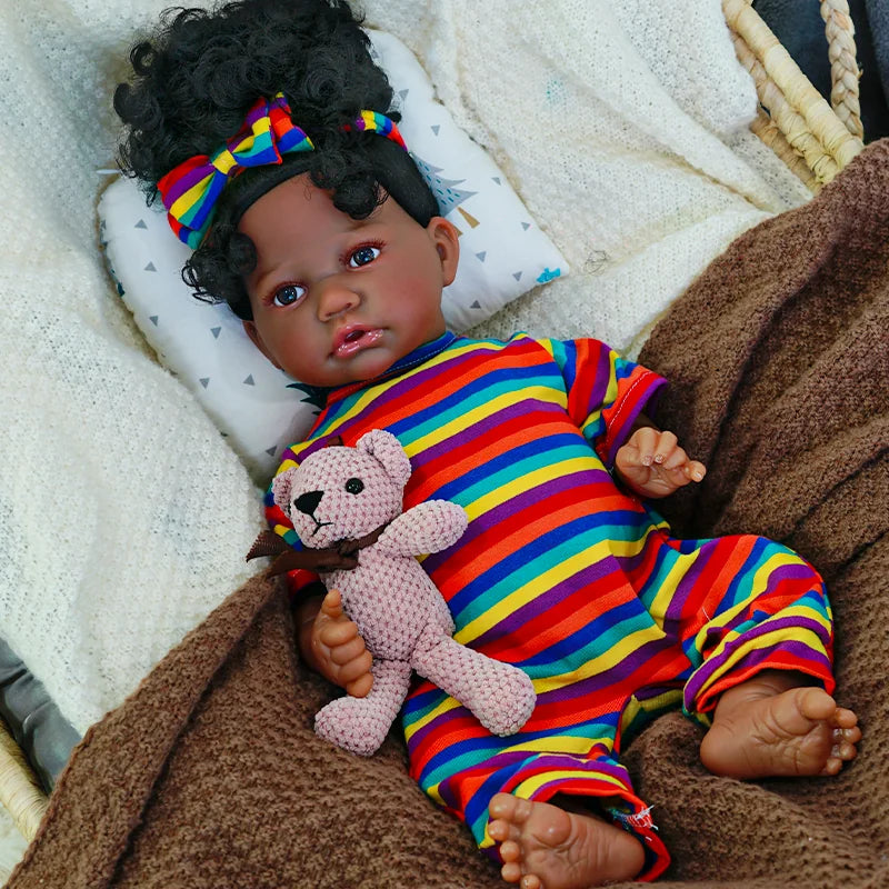 The first image shows a lifelike baby doll dressed in a rainbow-striped onesie, lying on a blanket, holding a small teddy bear.
