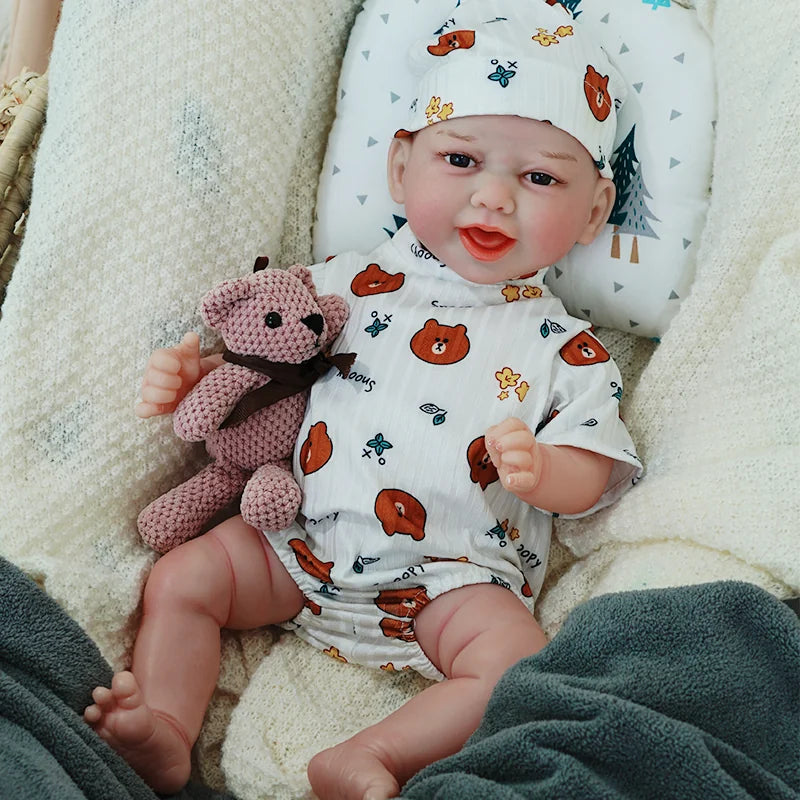 A smiling reborn baby doll dressed in a white onesie with animal prints, wearing a matching hat, and holding a knitted teddy bear.