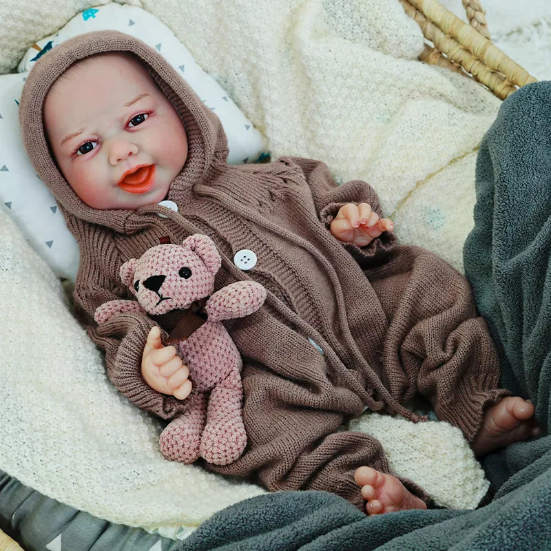 Reborn baby doll lying down in a cozy brown knit onesie, holding a knitted teddy bear, and smiling warmly.