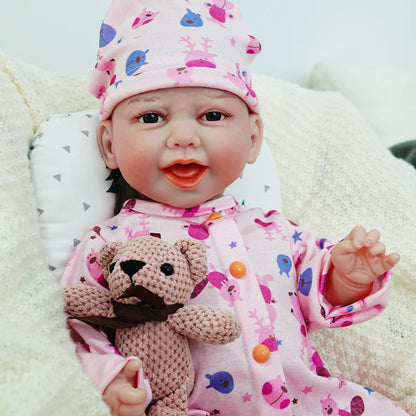 A smiling reborn baby doll wearing a pink outfit with animal prints and a matching hat, holding a knitted teddy bear.