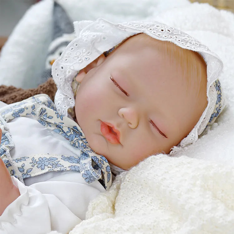 A realistic reborn doll peacefully sleeping, dressed in a white and blue floral outfit with a lace bonnet. The doll is nestled in a cozy, soft blanket.