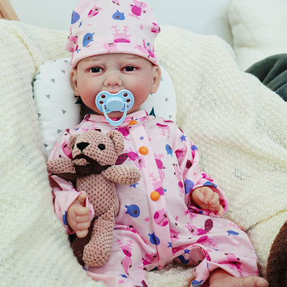 The reborn baby doll with a pacifier in its mouth, dressed in a pink animal print onesie, sitting with its knitted teddy bear.