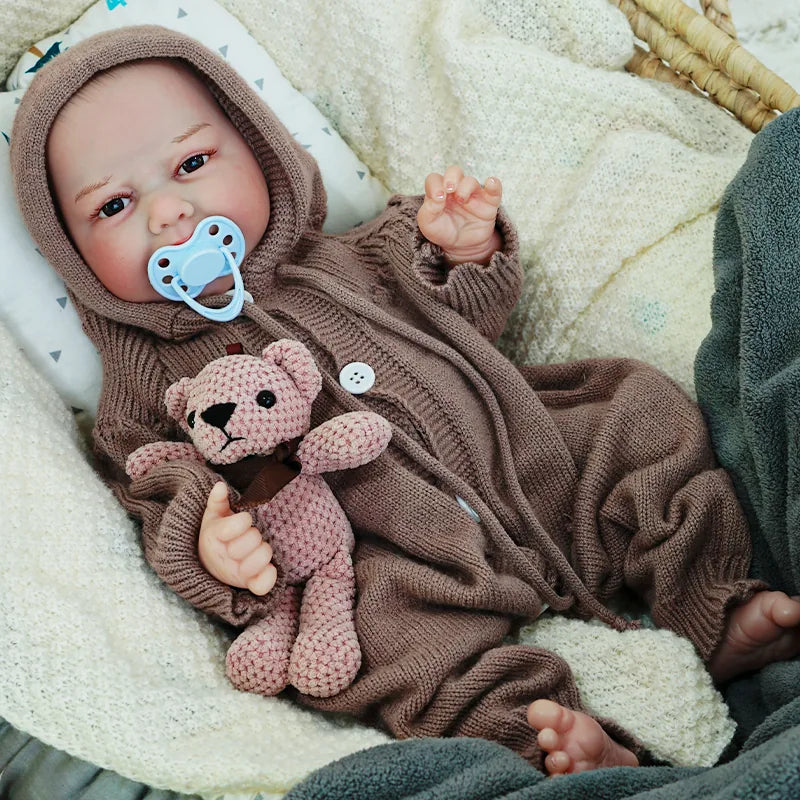 The reborn baby doll with a pacifier, dressed in a brown knit outfit, holding a soft teddy bear while lying on a blanket.