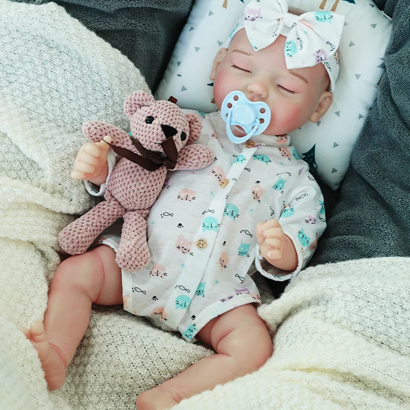 The reborn baby doll with a pacifier in its mouth, dressed in a blue patterned onesie and bow headband, cuddling with a knitted teddy bear.