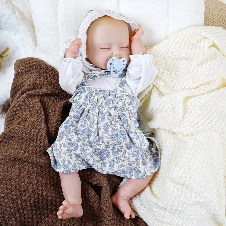A lifelike reborn doll dressed in a white blouse with blue floral overalls and a matching lace bonnet, holding a pacifier in its mouth and resting next to a stuffed bear toy on a cozy blanket