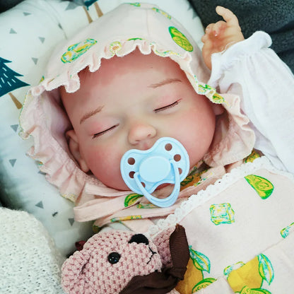 A peaceful reborn baby doll dressed in a green-patterned onesie, lying on a soft surface with a small teddy bear.