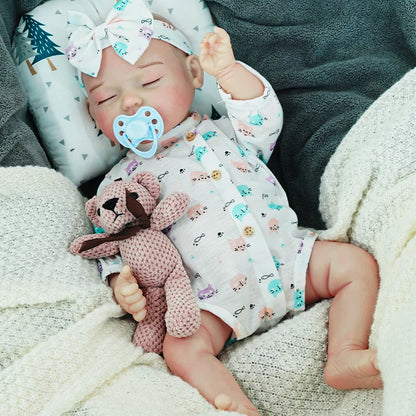 The reborn baby doll sleeping peacefully with a pacifier, dressed in a blue-patterned outfit, and holding a small knitted teddy bear.