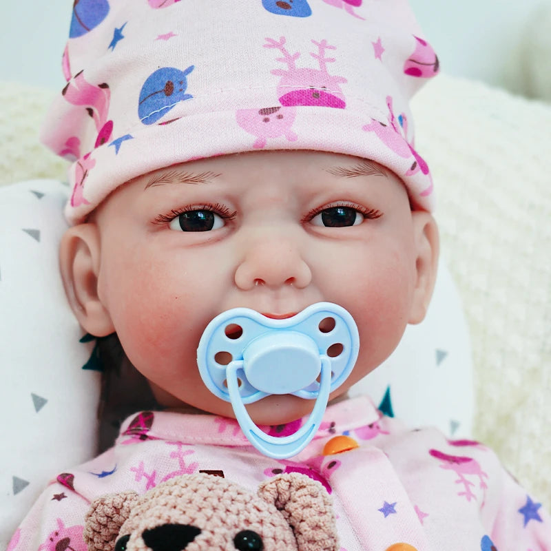 Close-up of the reborn baby doll’s face, wearing a pink outfit with a pacifier in its mouth and holding a knitted teddy bear.