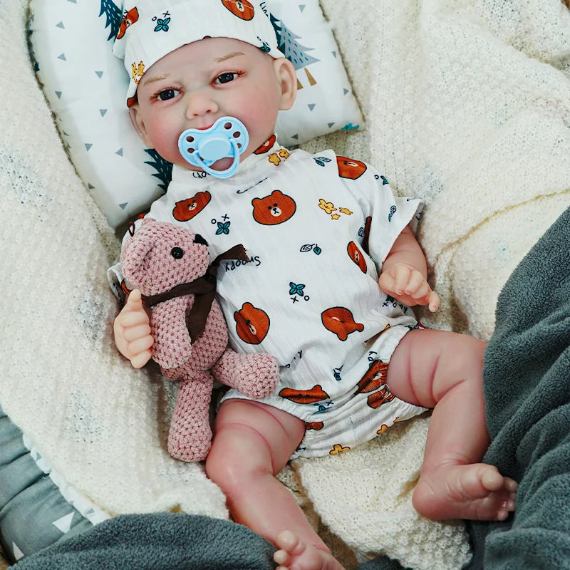 The reborn baby doll with a pacifier in its mouth, wearing a white onesie with animal prints, holding a teddy bear while sitting on a blanket.