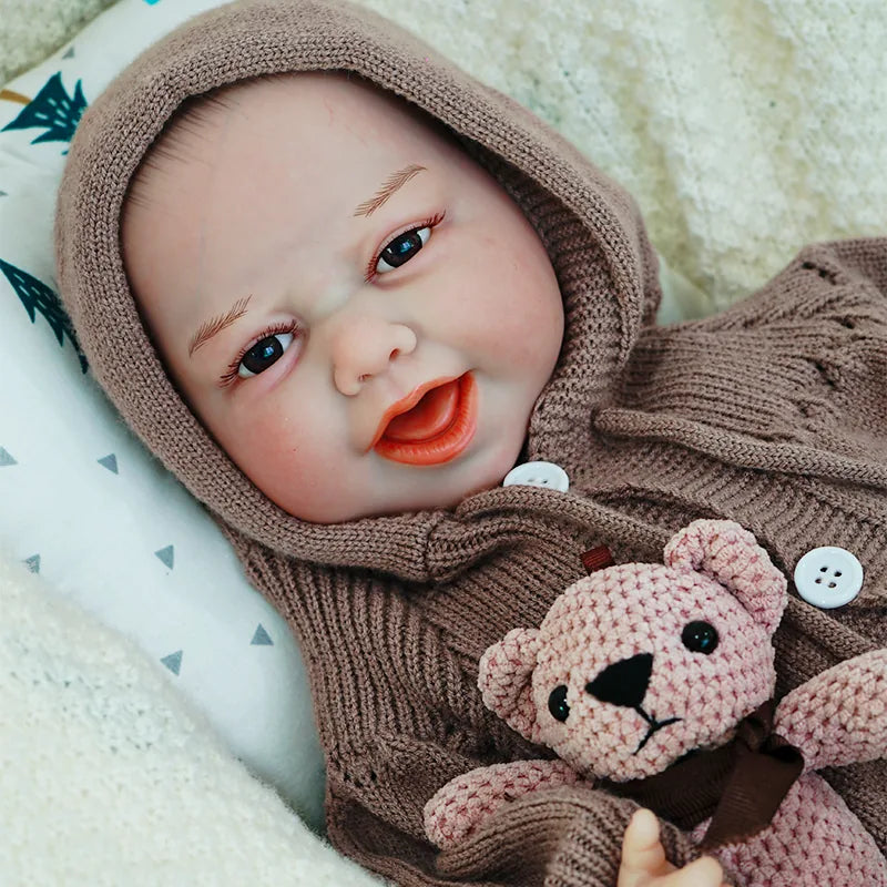 The reborn baby doll smiling happily, dressed in a brown onesie with a hood, and holding a knitted teddy bear.