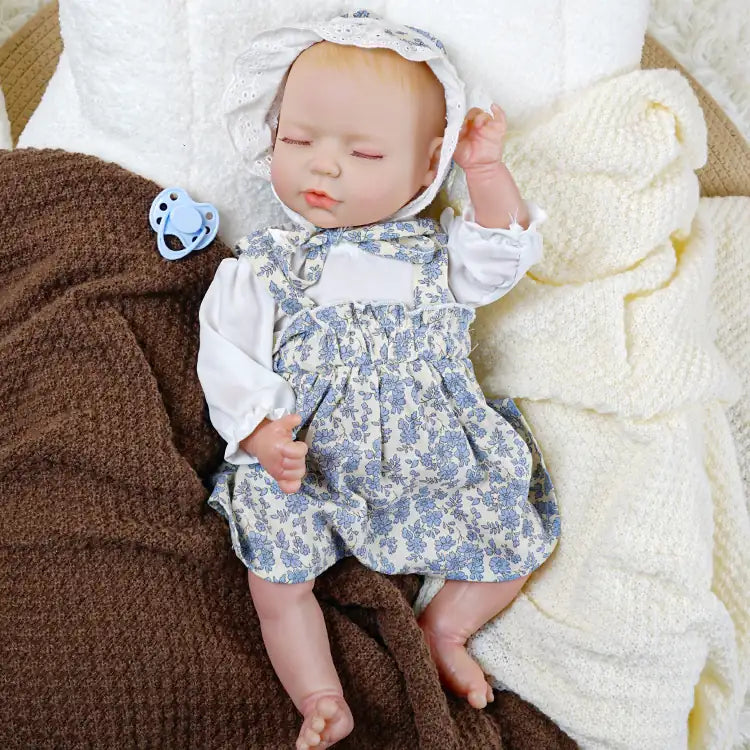 A lifelike reborn doll dressed in a white blouse with blue floral overalls and a lace bonnet, lying on a cozy blanket with a blue pacifier placed beside it