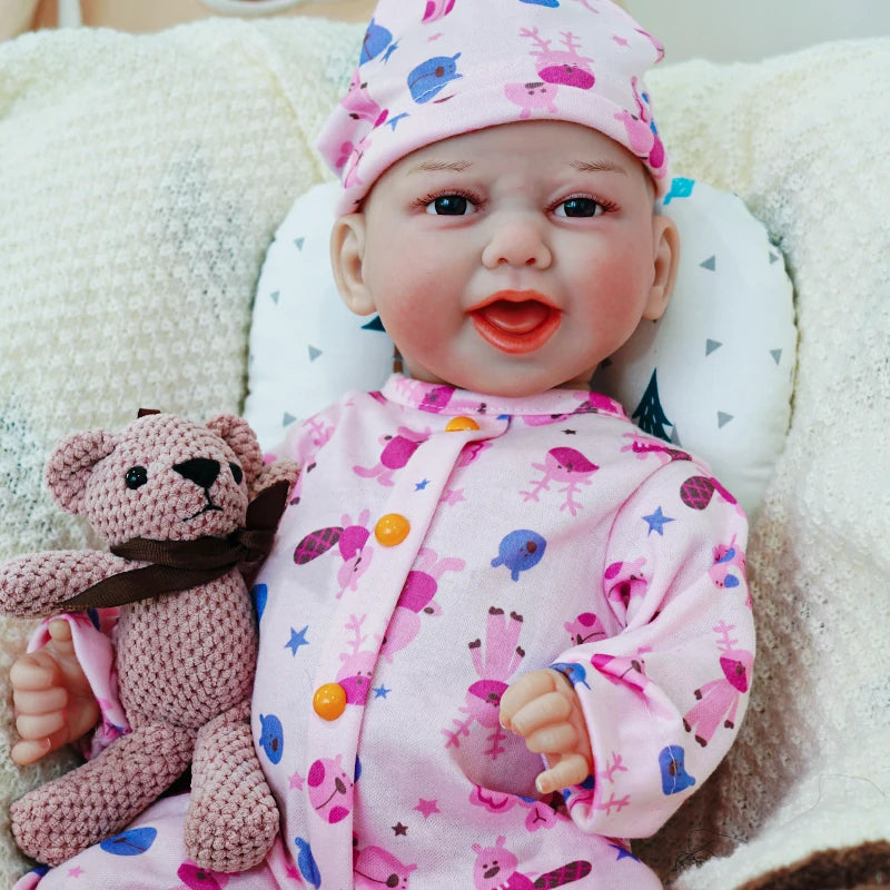 The reborn baby doll smiling, dressed in a pink animal print onesie and hat, with a knitted teddy bear in its hand.