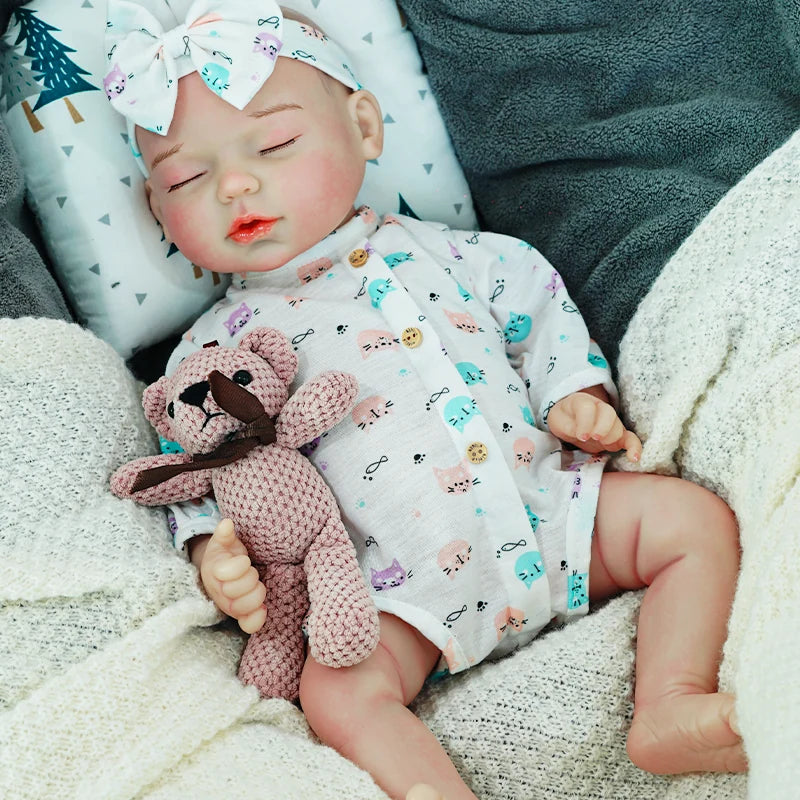  A close-up of the reborn baby doll sleeping, wearing a light onesie with blue accents and a bow headband, clutching a knitted bear.