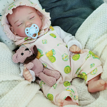 A close-up of a sleeping reborn baby doll dressed in a light green patterned outfit, lying on a soft blanket, with a knitted teddy bear beside it.