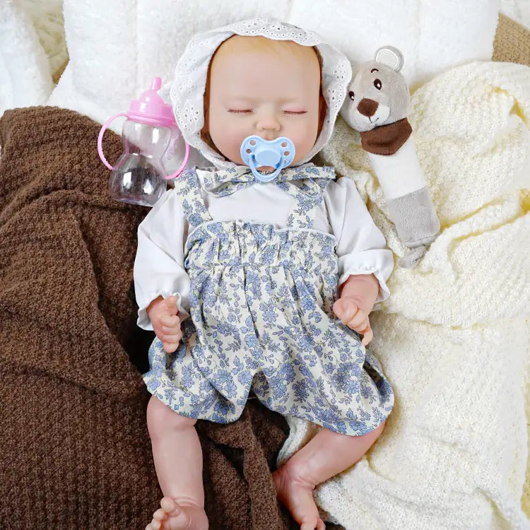 A lifelike reborn doll dressed in a white blouse with blue floral overalls and a lace bonnet, holding a blue pacifier in its mouth and raising one hand.