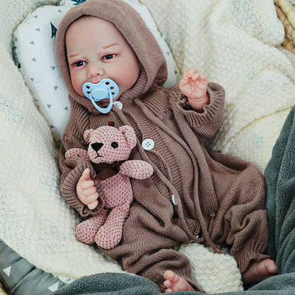 The reborn baby doll sitting up with a pacifier, dressed in a brown knit outfit, and holding a knitted teddy bear.