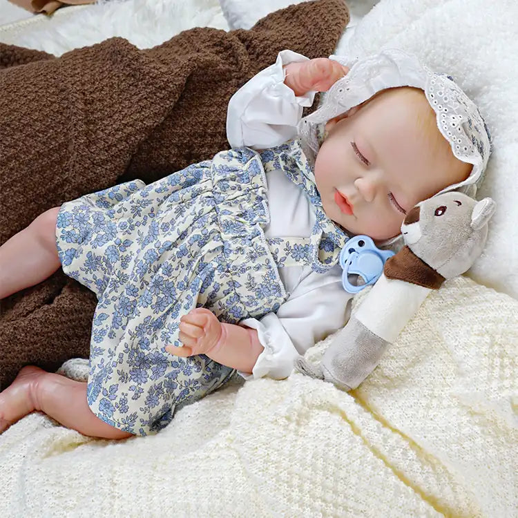 A lifelike reborn doll dressed in a white blouse with blue floral overalls and a lace bonnet, with eyes closed and hands gently raised.